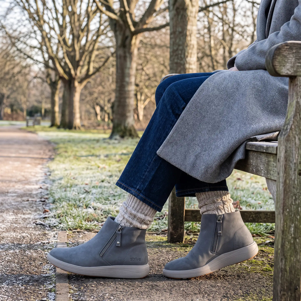 Woman wearing Strive Mayfair grey ankle boots with side zips while sitting on a park bench.