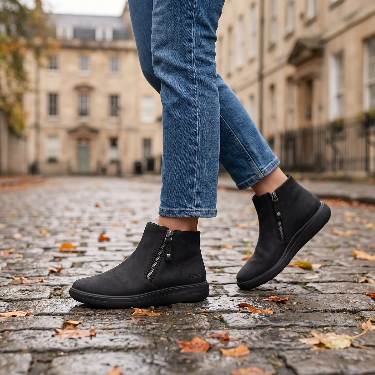 Woman wearing Strive Mayfair black ankle boots with side zips on an autumn cobblestone street.

