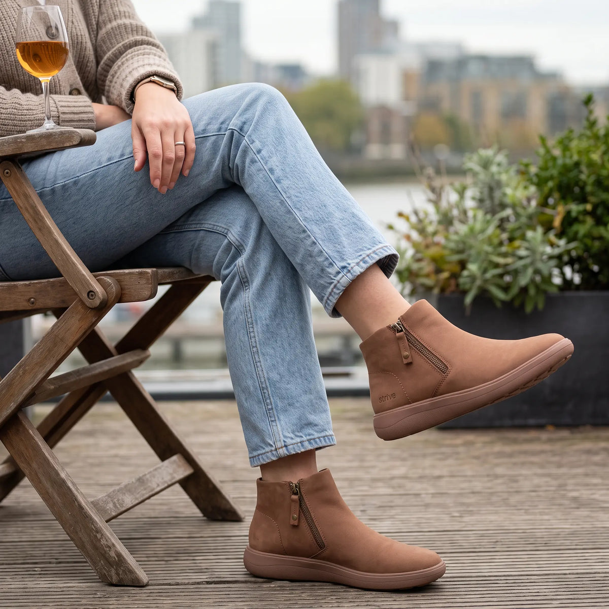 Woman wearing Strive Mayfair tan ankle boots with side zips while seated on an outdoor terrace.