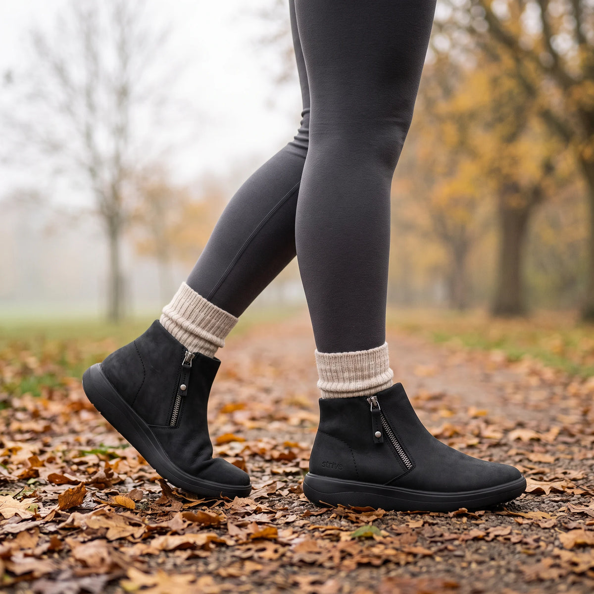 Black ankle boots with side zippers, worn with dark gray leggings and cream socks on a leaf-covered path in an autumn park.