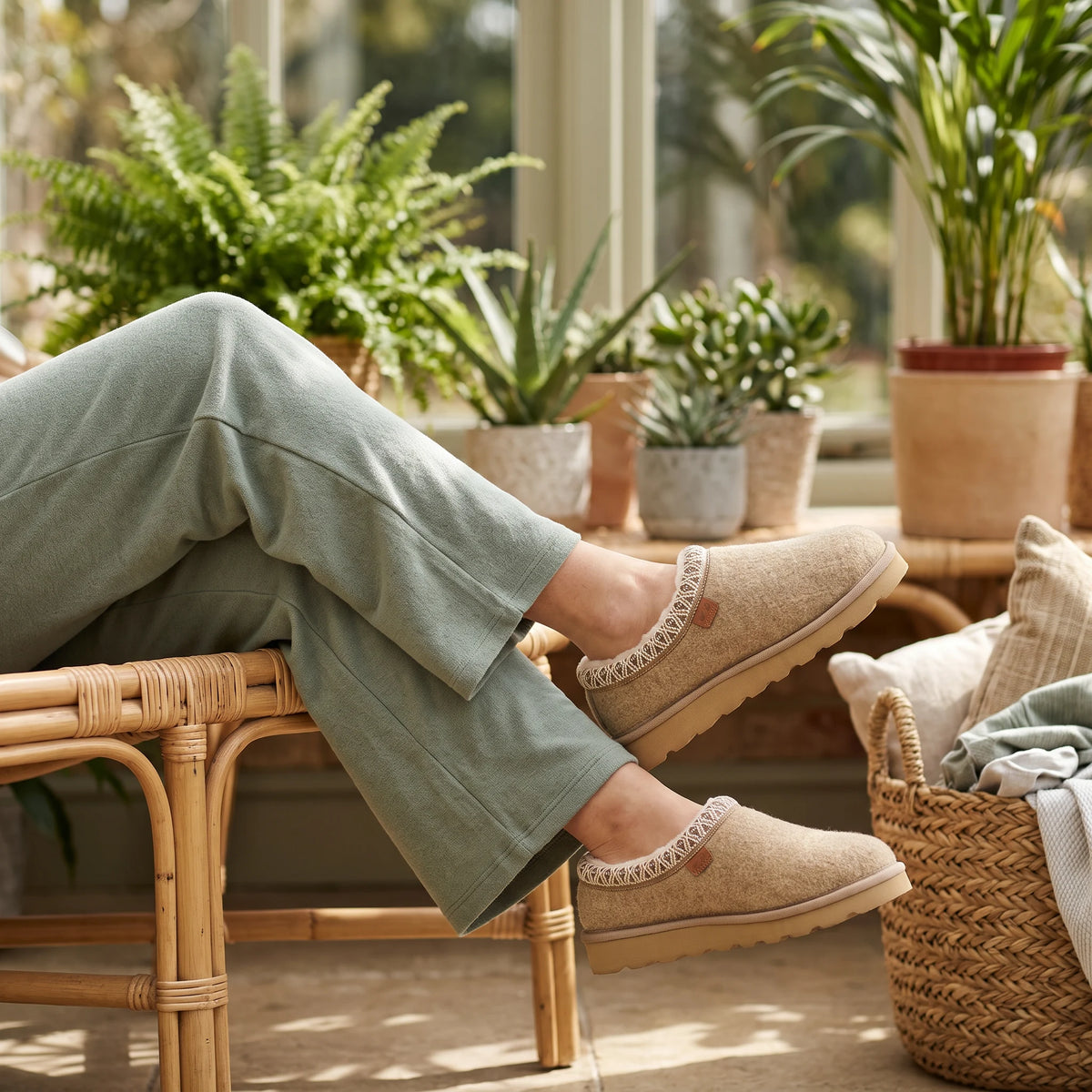 A person wearing beige slip-on slippers with white lace trim and sage green lounge pants, sitting on a rattan chair in a sunlit room with potted plants and a woven basket.