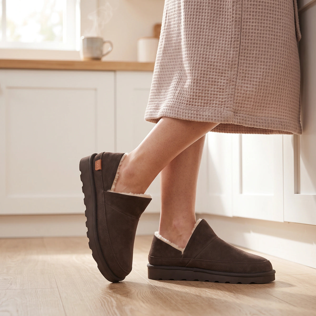 Brown Strive Alaska slippers with soft cream lining, worn in a bright kitchen.