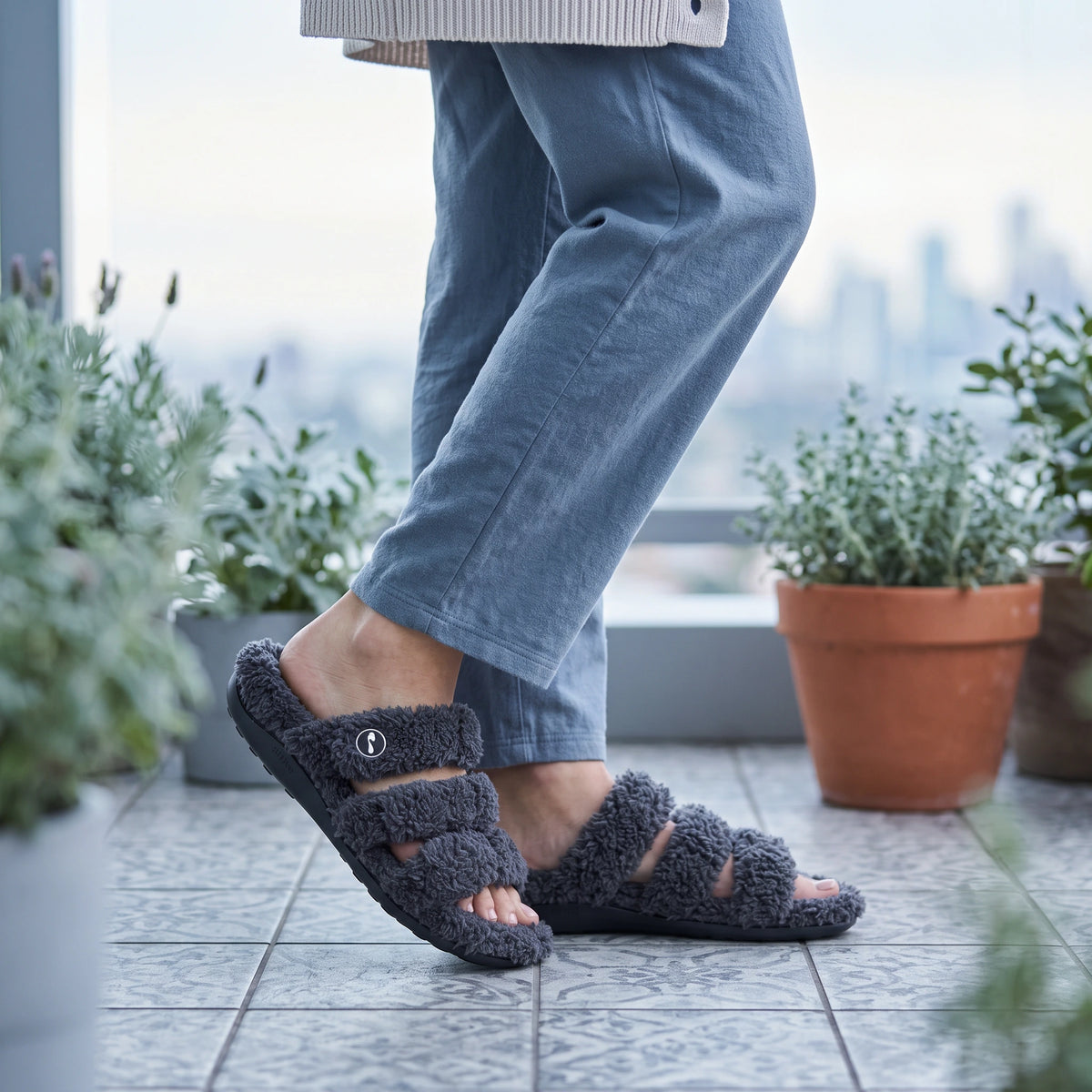 Grey Strive Zella teddy multi-strap slippers worn on a tiled balcony surrounded by plants.