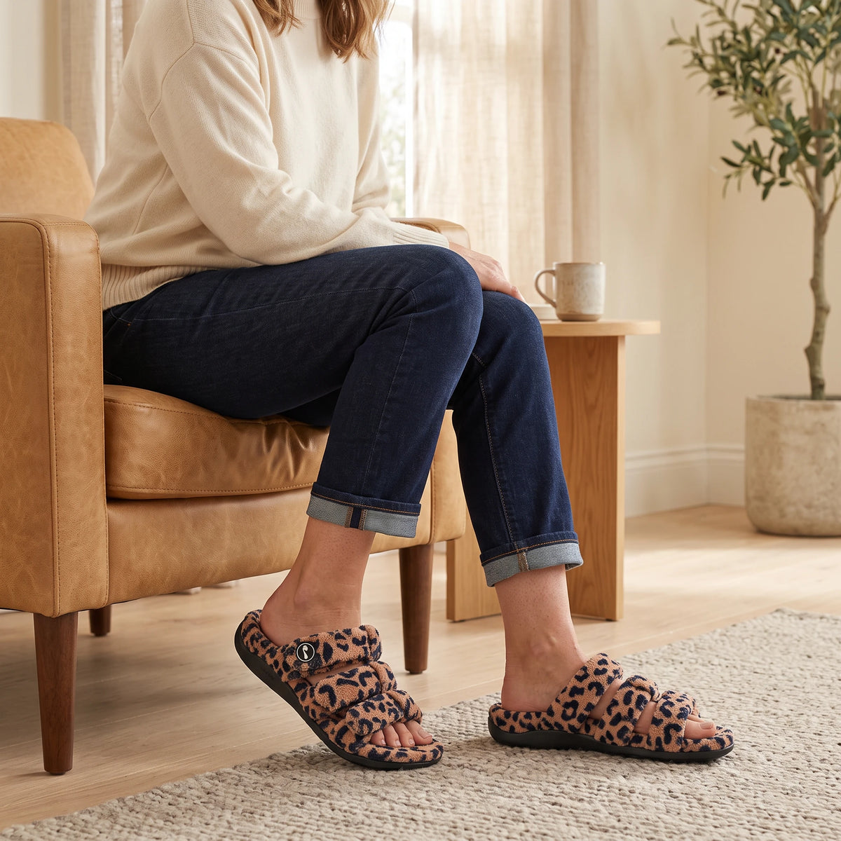 Leopard-print Strive Zella teddy multi-strap slippers worn while sitting in a bright living room.