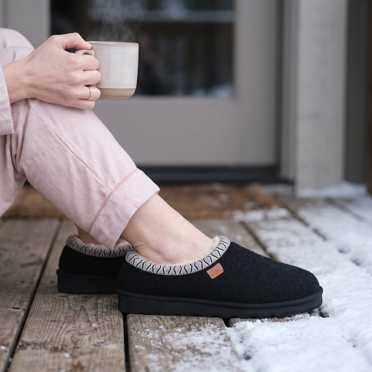 Black Strive Zurich felt slippers with patterned trim, worn on a snowy wooden deck.