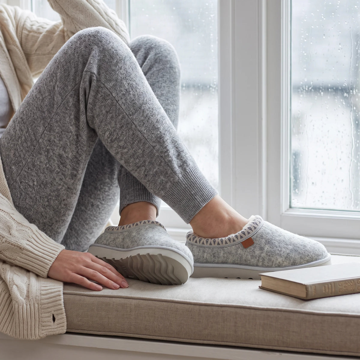 A person sitting by a window wearing light gray knit lounge pants and matching slip-on slippers with a small brown accent tab, next to a beige knit cardigan and a book on a cushioned window seat.